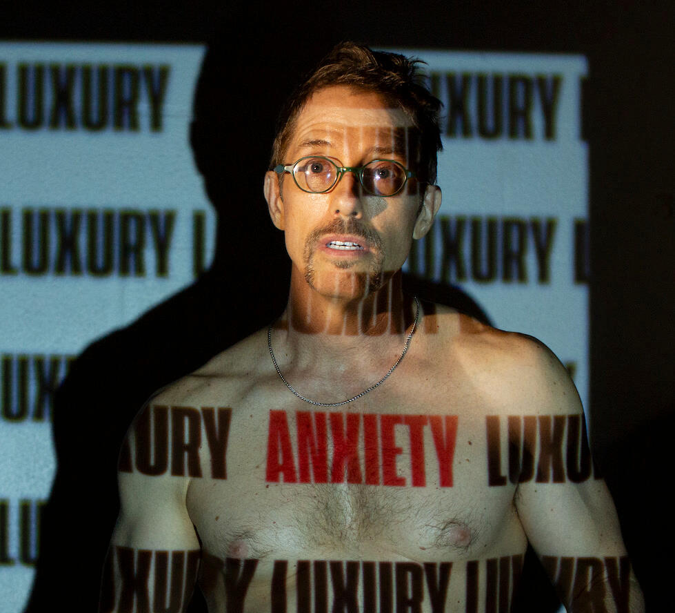 sirdanpaul shirtless against a wall of projected "LUXURY ANXIETY" text, photo by Taylor Cullen